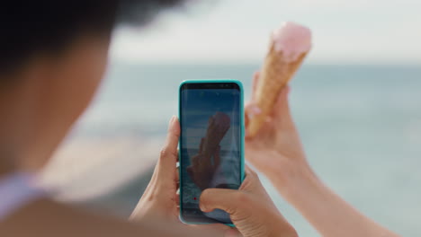 mujer joven sosteniendo un helado en la playa con un amigo tomando fotos usando un teléfono inteligente compartiendo un delicioso postre en las redes sociales 4k