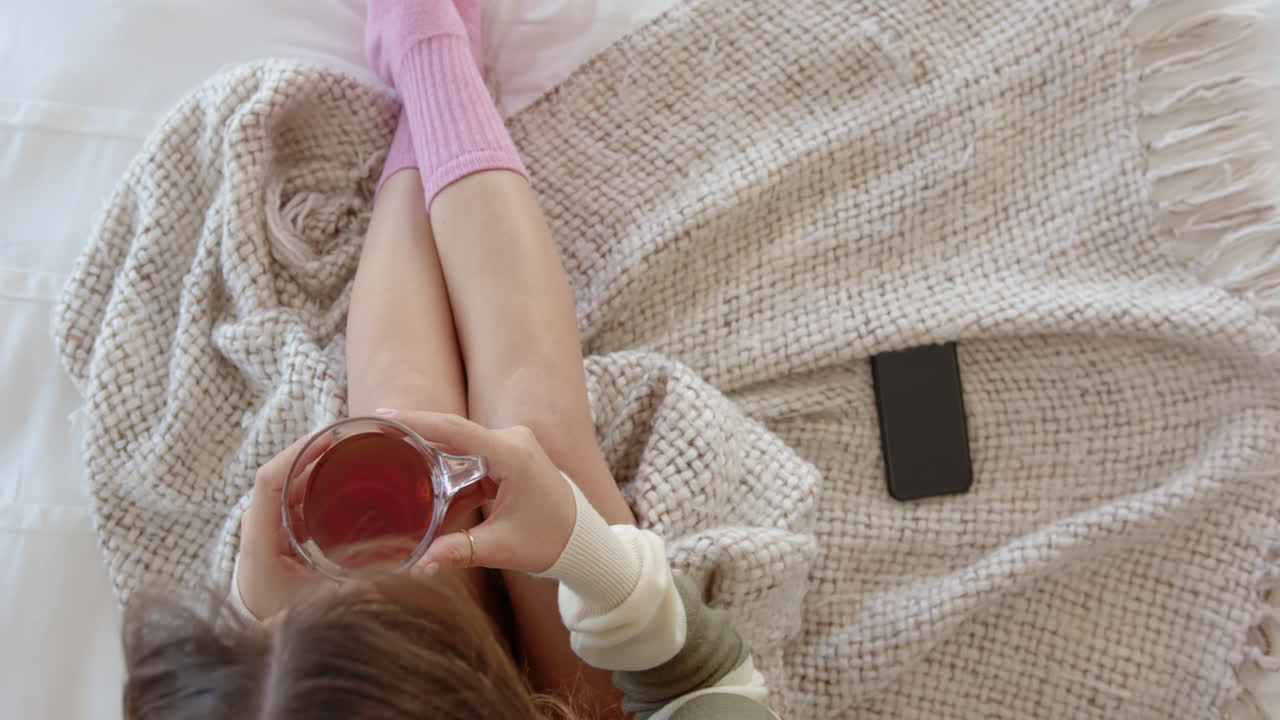 Relaxing in bed, woman drinking tea with smartphone beside her, copy space