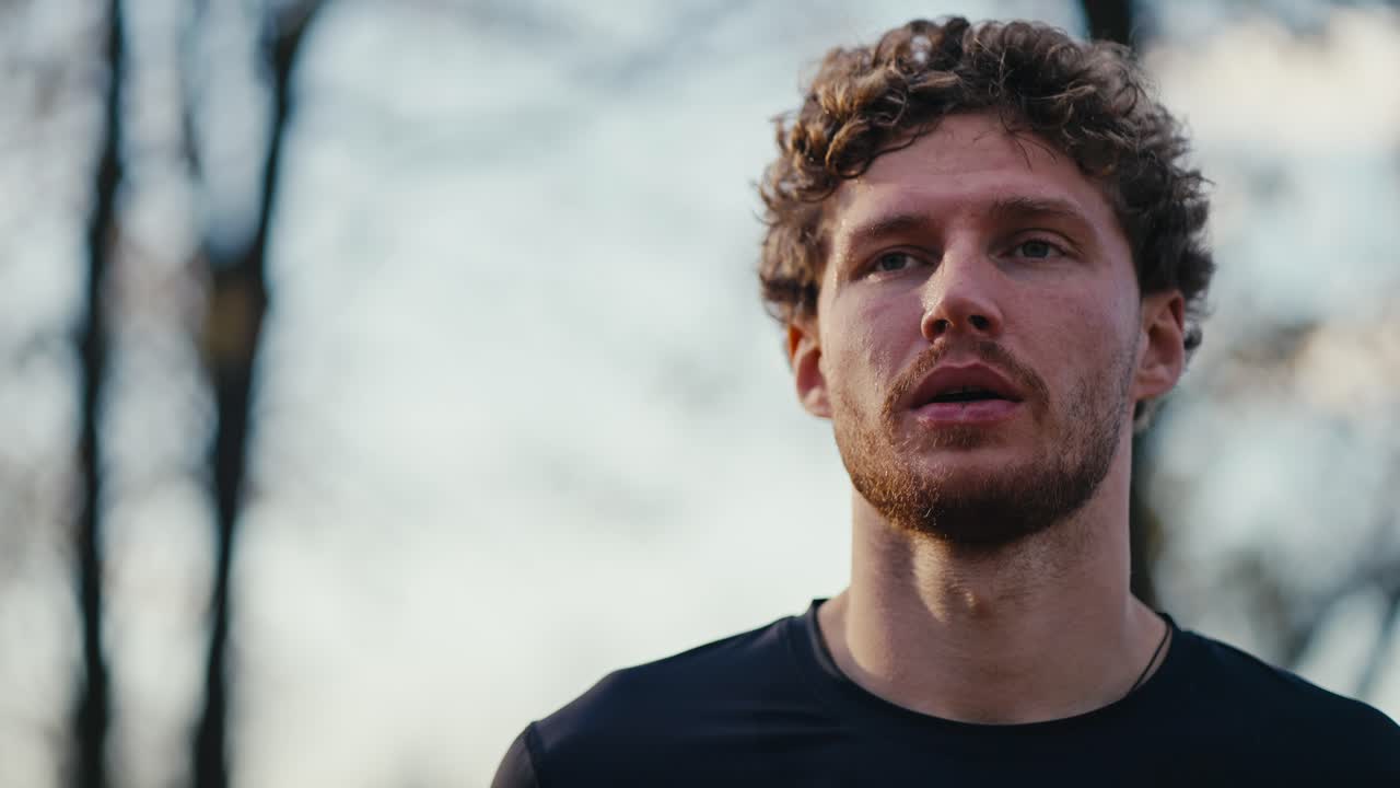 Portrait of a man with curly hair and a beard in a black sports uniform who wipes sweat from his face after a morning jog in the forest