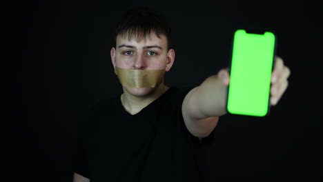 teenager with tape over mouth showing smartphone with green screen, censorship or cyberbullying concept