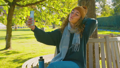 mujer sentada en un banco en el parque de otoño posando para una selfie en el teléfono móvil para publicar en las redes sociales