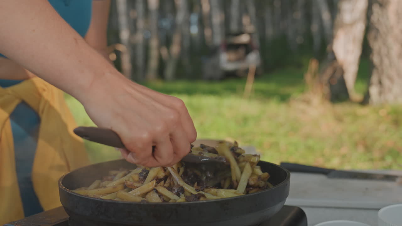 Camping meal with steaming beans and forest view. Black skillet on picnic table with woman cooking outdoors. Sunset scene of woman roasting beans in skillet beside forest and picnic setup