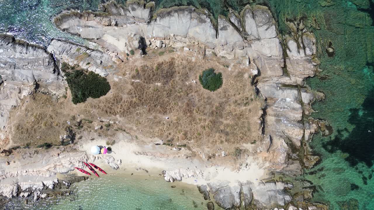 Droneutsikt ovenfra av en liten Drenia-øy nær Ouranoupoli, med steinete kystlinje, tørr sommervann og krystallklart turkist vann i en rolig lagune