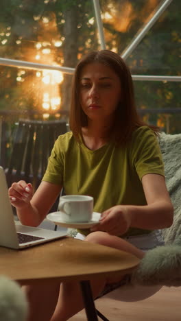 busy woman drinks tea in cup working in online sphere in tourist house surrounded by forest. business trip to another country with incredible views at sunset slow motion