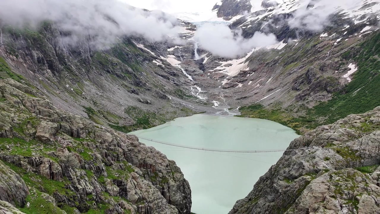 Turkise Triftsee-sjøen og hengebroen for fotgjengere omgitt av sveitsiske alper, skyer og en isbre-sjø, Triftbrücke, Triftgletscher, Bern, Sveits, utzoomende luftopptak