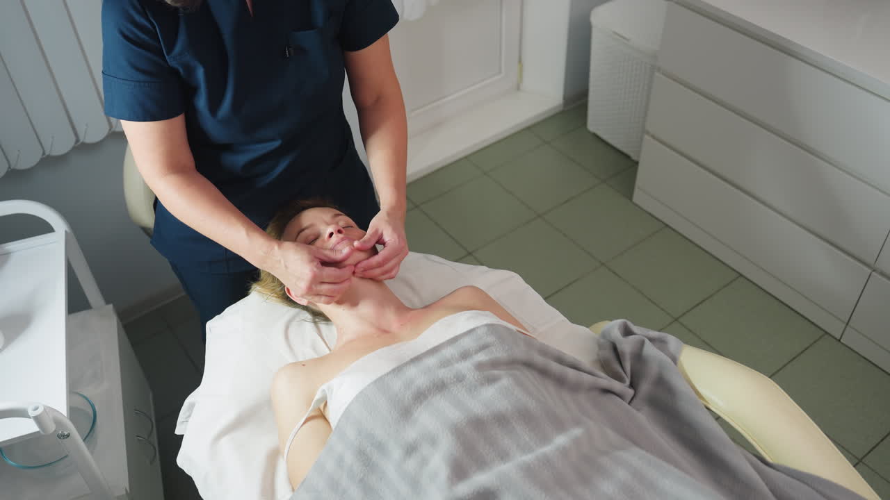 High angle of beauty expert smiling while cradling client jawline with gentle fingertips during relaxing massage session on soft linens in bright clinical room with organized product trolley nearby