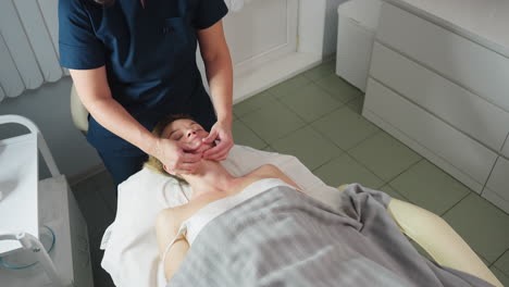 high angle of beauty expert smiling while cradling client jawline with gentle fingertips during relaxing massage session on soft linens in bright clinical room with organized product trolley nearby