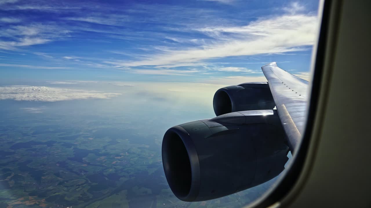 Looking through the window of a passenger airliner with the engines and wing in the foreground against a blue sky with clouds.