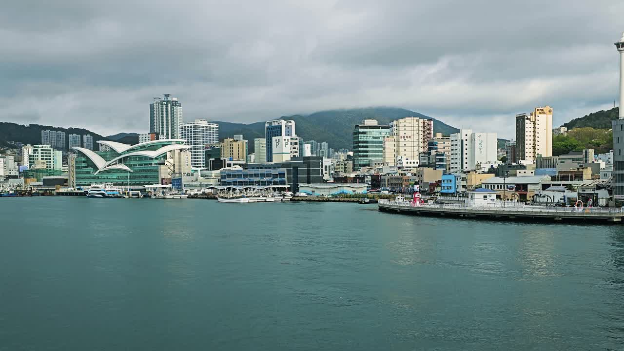 Panoramic view of Busan Tower and modern skyline in Jung District, Busan, with waterfront and city buildings under cloudy skies. Wide shot