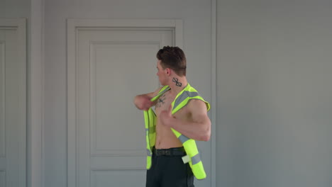 shirtless fair skinned young man stands before mirror and door sliding neon yellow jacket over shoulders in expressive dance moment highlighting neck and chest tattoos inside bright modern studio