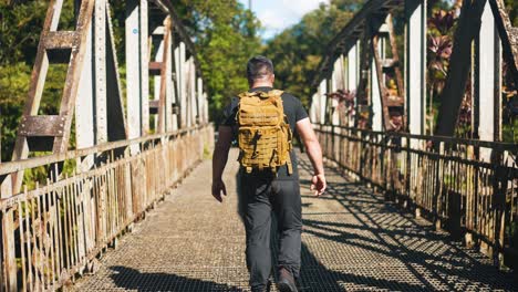 following young man with backpack walking on rusty old bridge outdoors on sunny summer day