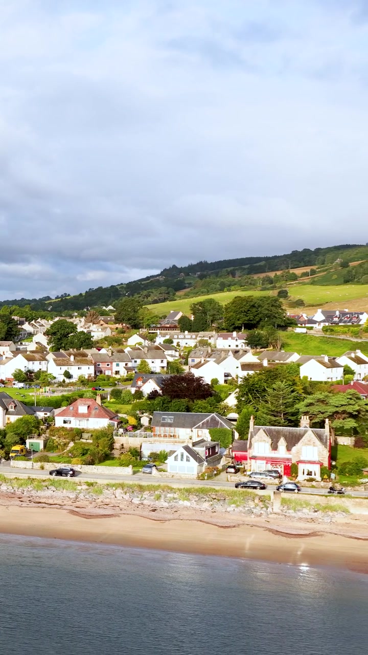 Câmera de drone faz uma panorâmica suave sobre uma vila costeira, revelando casas, colinas e litoral arenoso à luz do dia.
