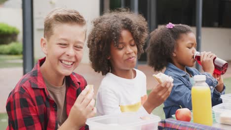 video of three diverse schoolchildren eating packed lunch and laughing in schoolyard