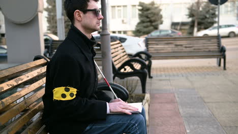 blind man with armband reading a braille book while sitting on bench outdoors