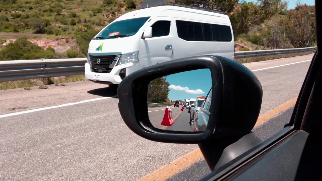 Specchietto laterale che riflette il traffico stradale, i coni di sicurezza e i veicoli durante un viaggio in autostrada con alberi a bordo strada e cielo blu a Huatulco, Oaxaca, Messico