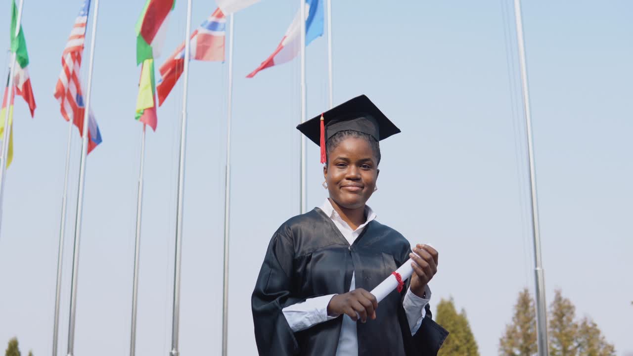 una joven afroamericana graduada está de pie frente a la cámara con un diploma en las manos. la estudiante está afuera con las banderas internacionales en el fondo.