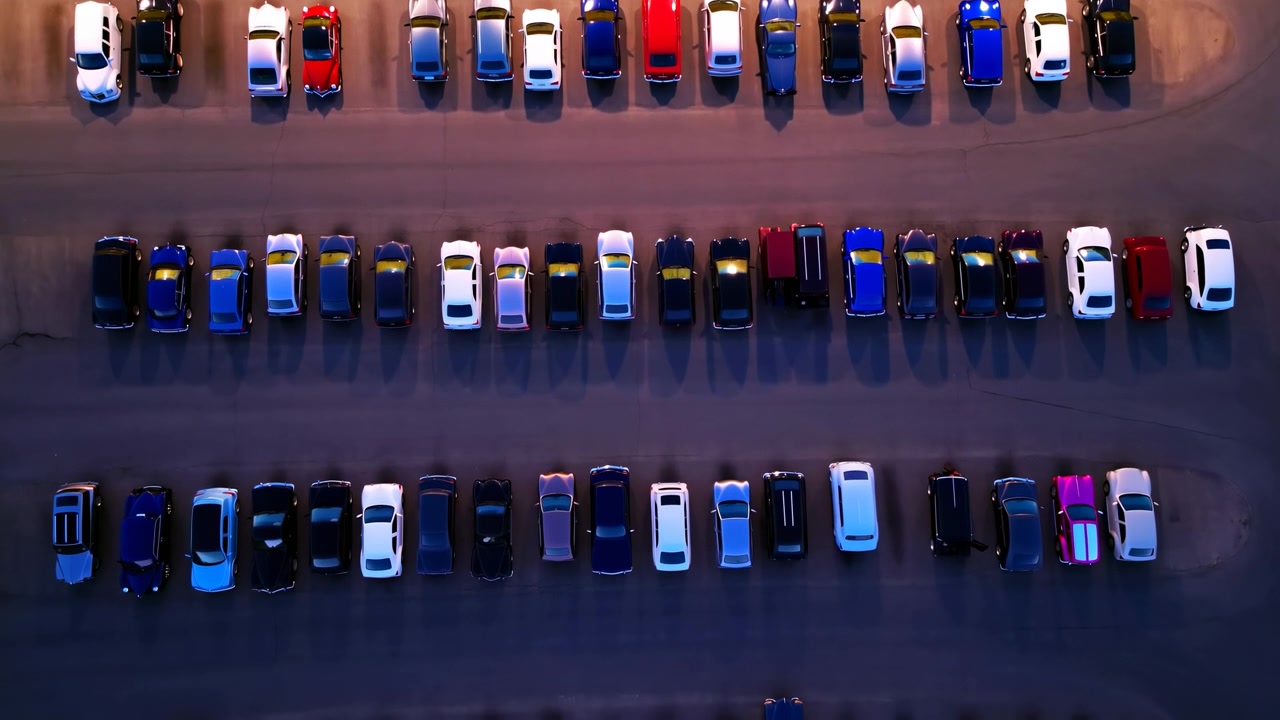 Top-down natkomposition, der viser tre parallelle rækker af parkerede biler på en oplyst parkeringsplads. Ovenlys skaber varme højdepunkter på køretøjernes tage og lange