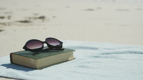 Close-up-of-book,-sunglasses-and-towel-on-beach,-in-slow-motion,-with-copy-space