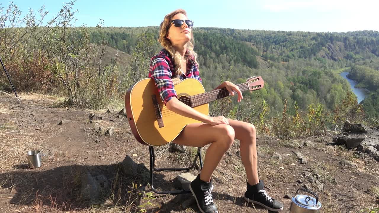 젊은 금발 여성 관광객은 기타와 함께 강 위의 절벽에 캠핑 의자에 앉아 태양에 basking