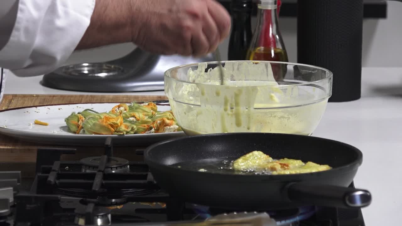 A chef prepares stuffed zucchini flowers by dipping them in batter and frying them in a pan. The kitchen is well-equipped, showcasing fresh ingredients and utensils