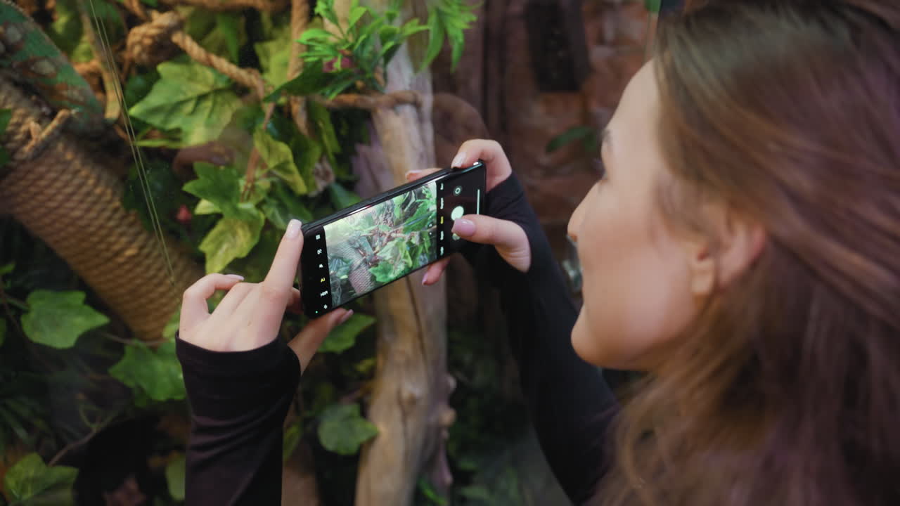 Woman with blond hair holds smartphone in landscape orientation, capturing vibrant indoor greenery through glass with focused expression, surrounded by lush leaves and natural textures