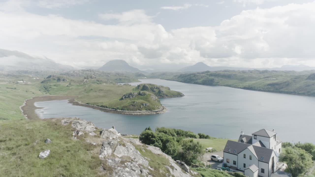 Slow drone panning shot of a mansion house in the foreground, a Loch in Scotland in the middle and tall mountains and clouds in the distance and sun shining
