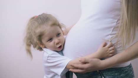 Hija-Abraza-El-Vientre-De-Mamá-Embarazada-De-Pie-En-La-Pared-De-La-Habitación