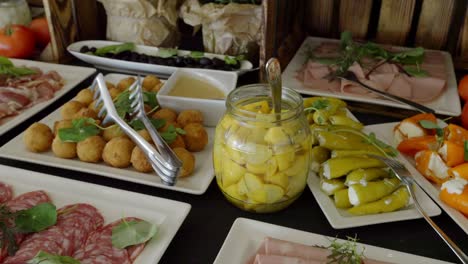 a table with snacks, on which there are plates of marinated bell peppers, cold cuts, pickles, tomatoes, and olives