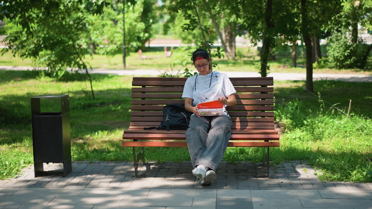 햇빛 아래 공원 벤치에 앉아 있는 남자, 모자와 백팩을 멘 젊은 남자가 휴식을 취하며 스트레칭을 하고, 주변을 둘러봅니다. 쓰레기통 옆 나무 벤치, 잎이 무성한 나무와 푸른 잔디, 얼룩덜룩한 그늘, 따뜻함