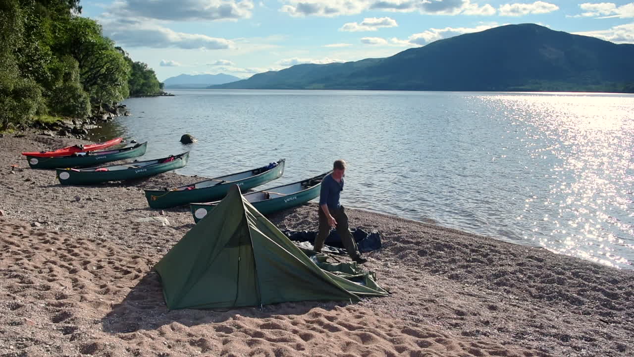 Man Setting Up A Tent Near The Boats On The Sandy Shore Of Caledonian Canal, Loch Ness, Scottish Highlands In Scotland On A Summer Day - Timelapse