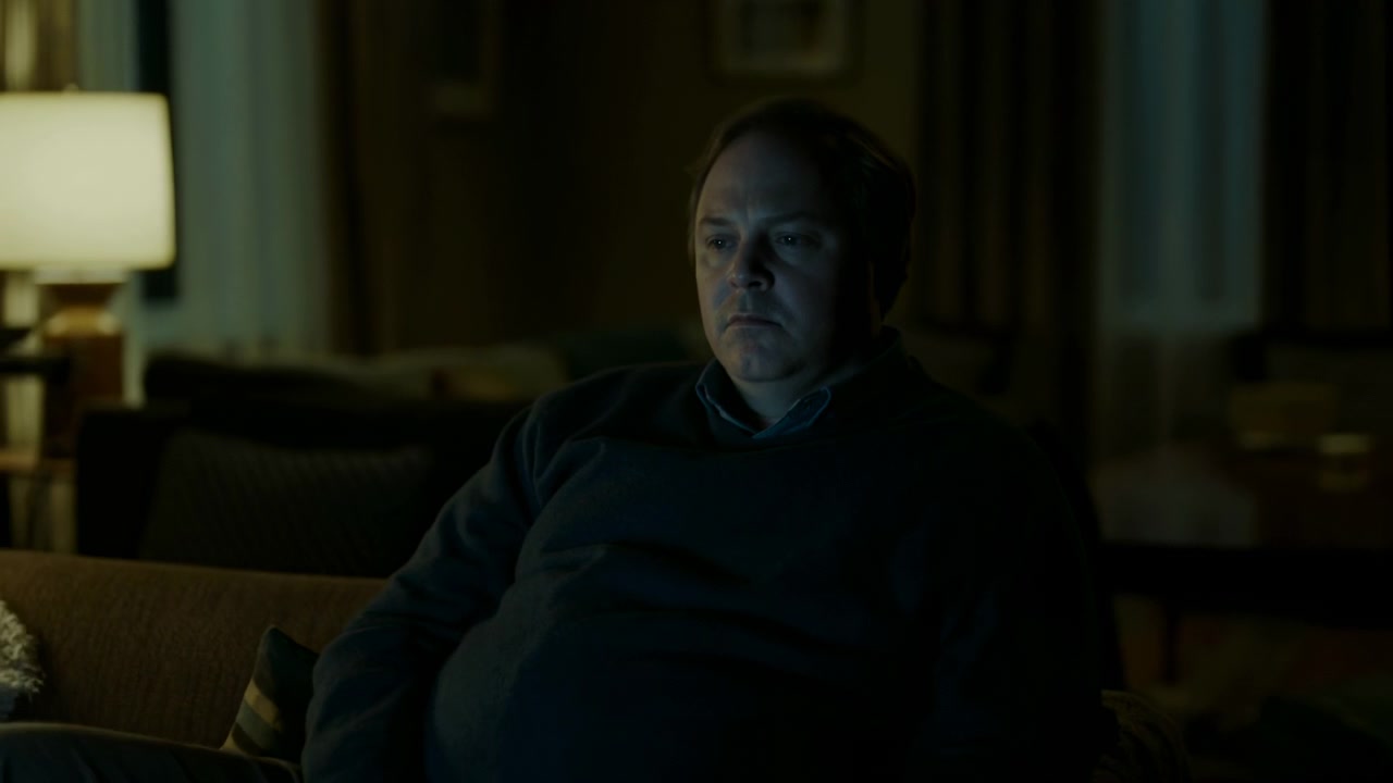 Seated man keeps his gaze fixed on monitor as framing gradually widens, revealing armchair, curtains, nearby furniture and dim interior with blue screen light across his face.