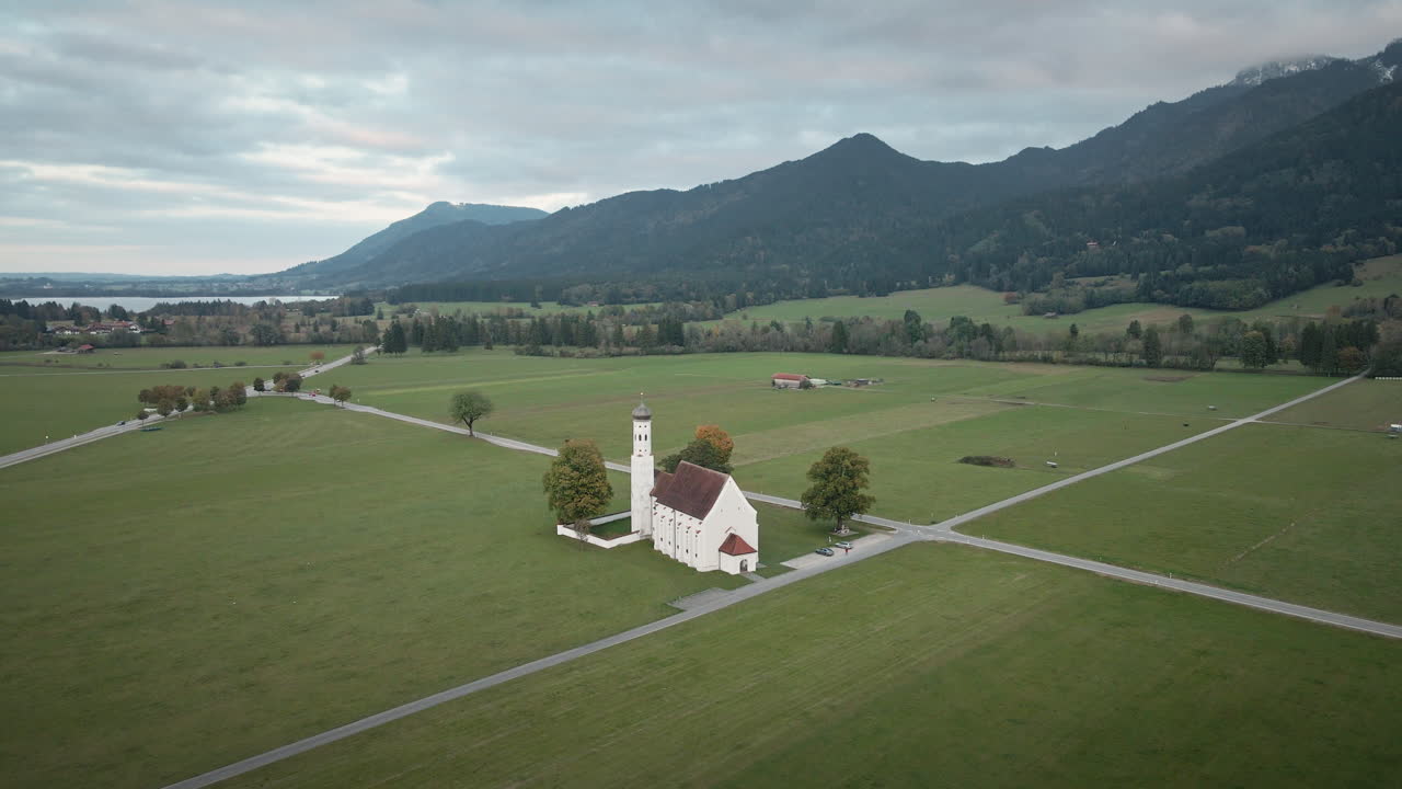 Tasainen 4K-droneveto Pyhän Colomanin kirkosta Schwangaussa, joka laajenee näyttämään niittyjä, maalaisteitä, kaukaisia järviä ja Baijerin Alppeja pilvisen taivaan alla.