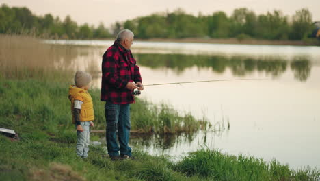 el anciano y su nieto están pescando juntos en la orilla de un hermoso lago por la mañana