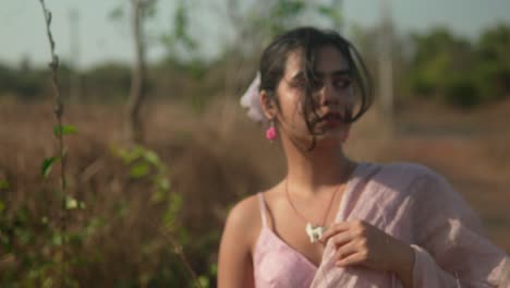 Young-woman-in-pink-dress-with-a-flower-in-hair-standing-in-a-field,-soft-focus,-natural-light