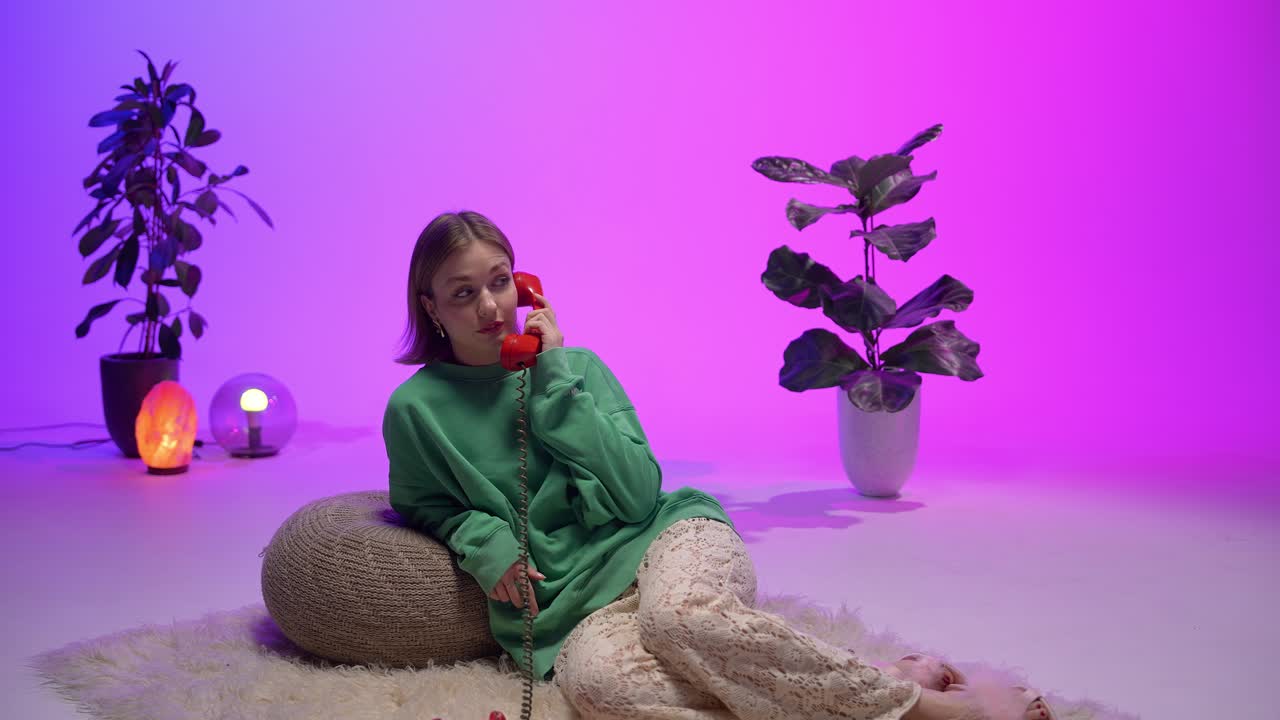 A stylish woman in a green sweater and lace pants gently hangs up a vintage red rotary phone while lounging on a fluffy rug in a cozy, modern-retro room with vivid ambient purple-pink lighting