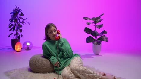 a stylish woman in a green sweater and lace pants gently hangs up a vintage red rotary phone while lounging on a fluffy rug in a cozy, modern-retro room with vivid ambient purple-pink lighting