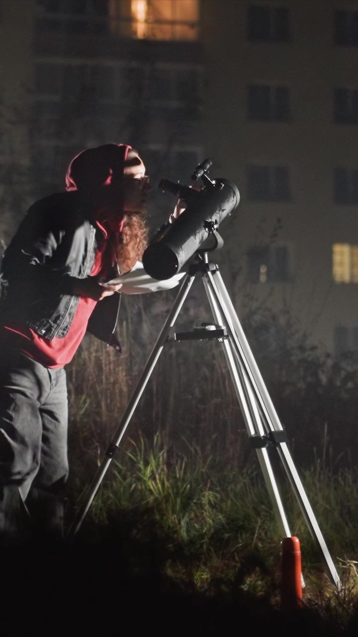 Krøllhåret person i rød hettegenser står nær et teleskop. Personen skriver notater med høyre arm nær det stativmonterte teleskopet. Scenekomposisjonen har opplyste vinduer på en fleretagers bygning.