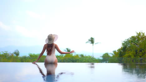 Vista-Trasera-De-Una-Mujer-Sexy-Solitaria-Sentada-En-El-Borde-De-La-Piscina-Infinita-Con-Una-Vista-Impresionante-De-La-Isla-Tropical-Y-El-Horizonte-Marino