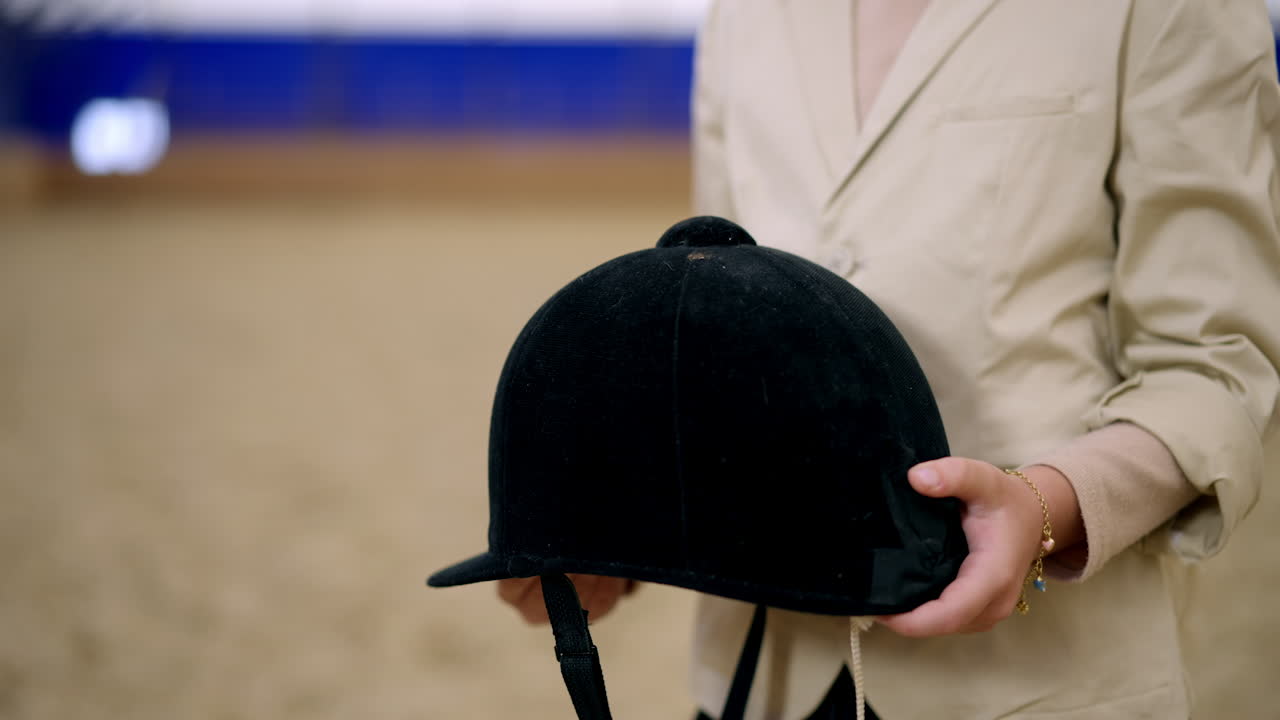 Beautiful young long-haired girl holding a black jockey helmet in her hands. Cute kid smiling to the camera standing still. Riding hall at backdrop in blur.