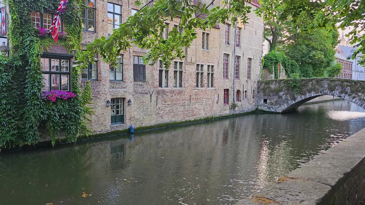 Malerisk kanal i den gamle bydel med murstenshuse, en stenbuebro, hængende blomster, efeu-beklædte mure, løvrige trægrene og roligt, reflekterende vand i en naturskøn europæisk ramme.
