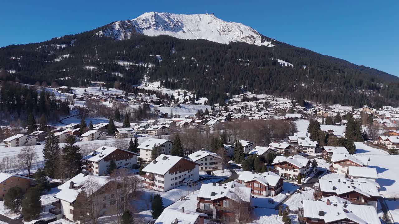 Veduta aerea di un villaggio svizzero innevato con case alpine e una drammatica cima montuosa che si innalza sopra la foresta sotto un limpido cielo blu invernale. Vista panoramica. Capanne di legno su un pendio innevato.