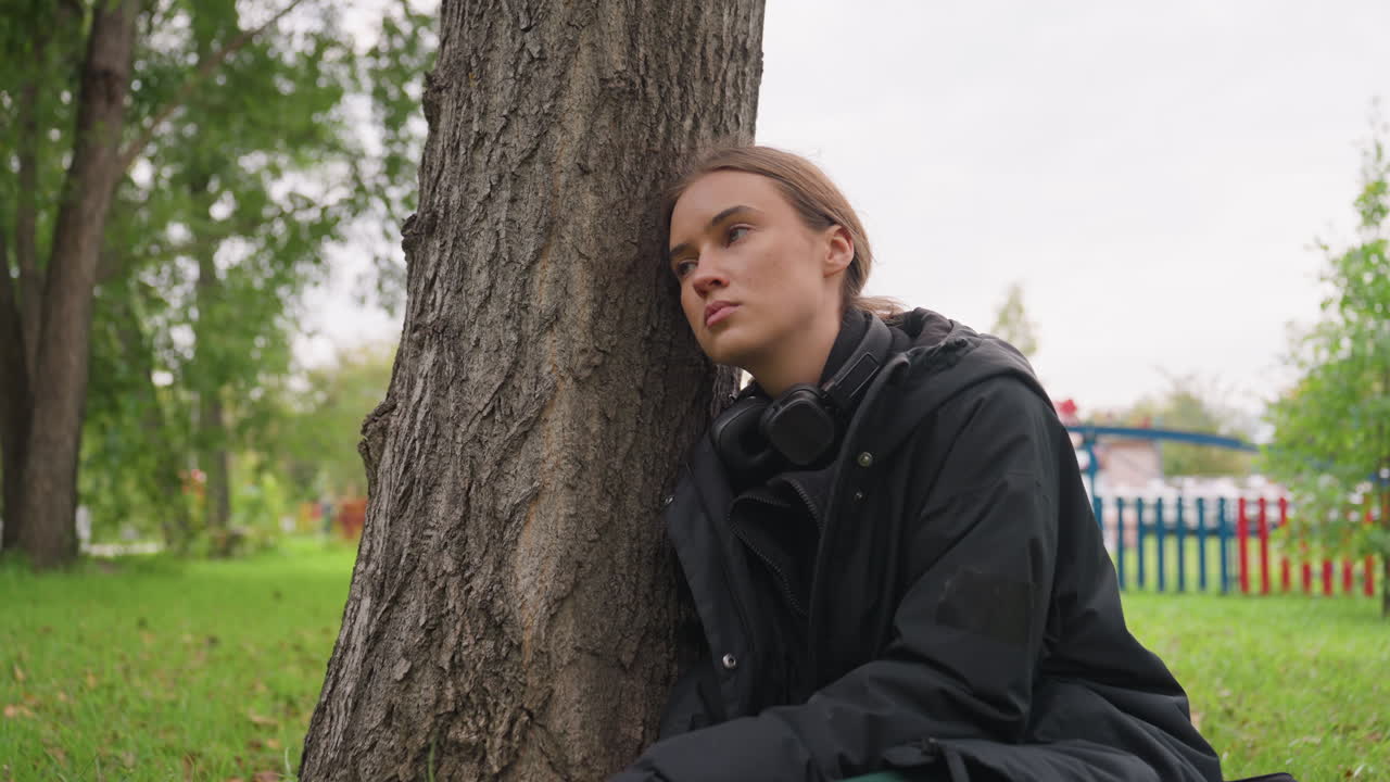 Junge Frau lehnt an Baum, nachdenkliche urbane Parksituation mit Kopfhörern und Jacke, grünes Gras und farbenfroher Zaun im Hintergrund. Nahaufnahme fängt einen introspektiven Blick und eine bewölkte Himmelsstimmung ein. Ausgedacht