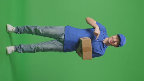 full body of asian male courier in blue uniform smiling and pointing to a carton in hand while delivering it on green screen background in the studio