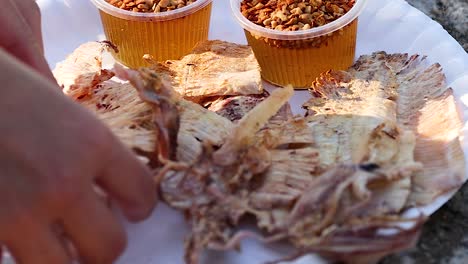 close-up of hands preparing grilled squid with spicy dipping sauce on a paper plate.