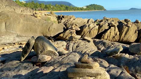 smooth rising shot over a rocky shoreline with many rock cairns piled up among the rocks, as the view opens out to a broad tropical and sunlit bay in the background