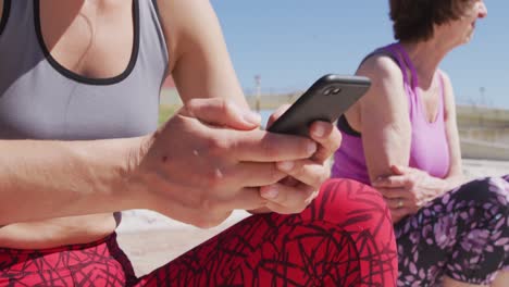 Kaukasische-Frauen,-Die-Sich-Hinsetzen,-Und-Eine-Frau,-Die-Ihr-Telefon-Am-Strand-Und-Vor-Blauem-Himmel-Im-Hintergrund-Benutzt