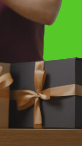 vertical video of man putting present in gift wrapped box decorated with ribbon into gift bag on table shot against green screen