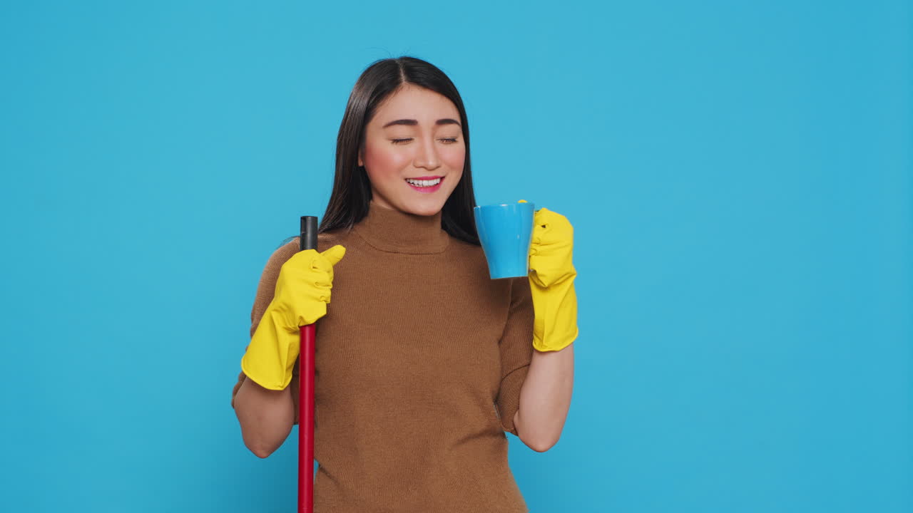 Cheerful housekepper enjoying cup of coffee while cleaning house with broom