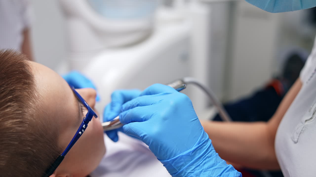 Hands in blue latex gloves take metal tool and drill machine to the boy's mouth. Teeth treatment in child dental cabinet. Close up. Blurred backdrop.
