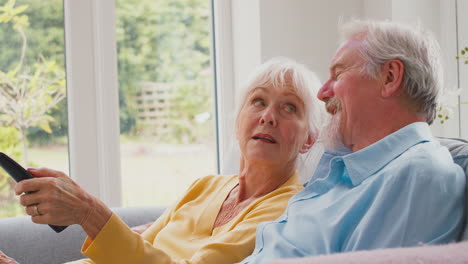 Pareja-De-Ancianos-Jubilados-Con-Control-Remoto-Sentados-En-Un-Sofá-En-Casa-Viendo-Televisión-Y-Riendo-Juntos